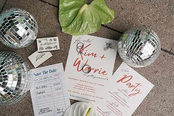 Invitation suite wedding invitation flat lay with RSVP and save the date cards, rings, pearl earrings, anthurium and disco balls on stone pavement