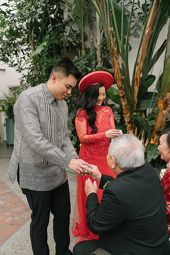 Tea ceremony moment as bride in red ao dai and groom serve tea cups to elders on a courtyard patio with tropical plants behind