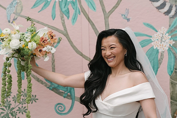 Bridal portrait of a laughing bride holding a bouquet in an off the shoulder dress with veil and drop earrings before a floral mural wall