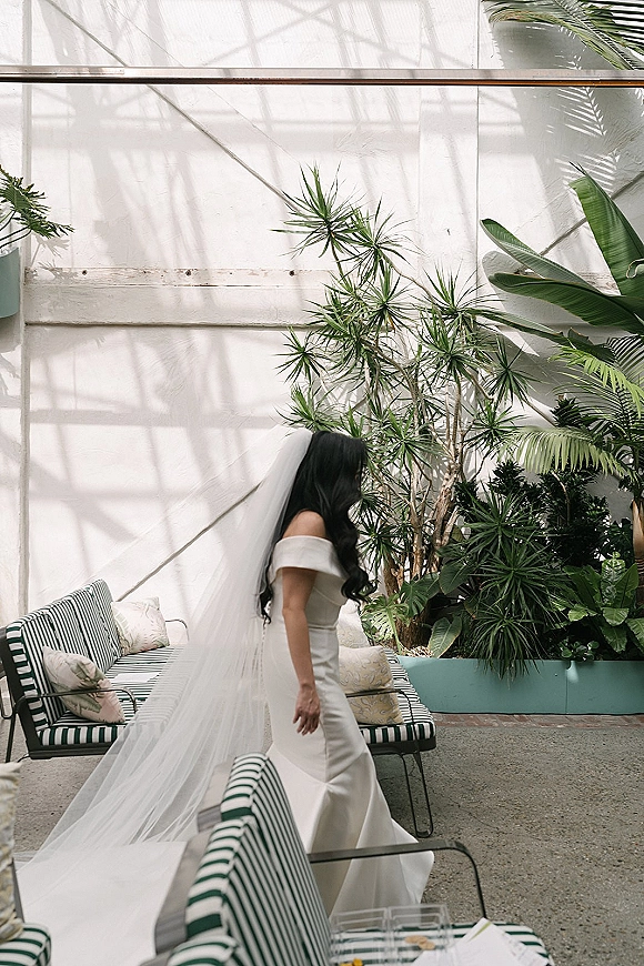 Bridal portrait of a bride in an off the shoulder gown with a long cathedral veil on a striped patio sofa under greenhouse shadows