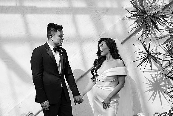 Couple portrait in a black and white wedding portrait style, bride in veil and dress holding hands with groom in tux by a stucco wall