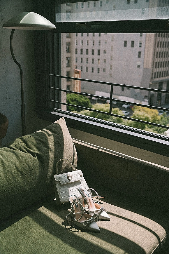 Bridal shoes in a wedding shoes flatlay with crystal slingback heels and white clutch on a sofa by a sunny window overlooking city buildings
