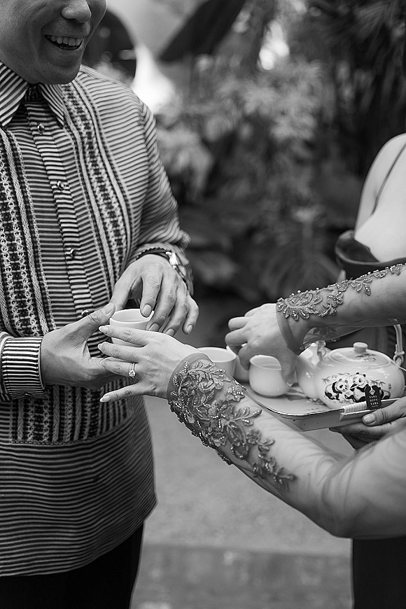 Wedding tea ceremony hands hold a teacup and pour from a porcelain teapot on a tray, lace sleeves and striped jacket amid greenery