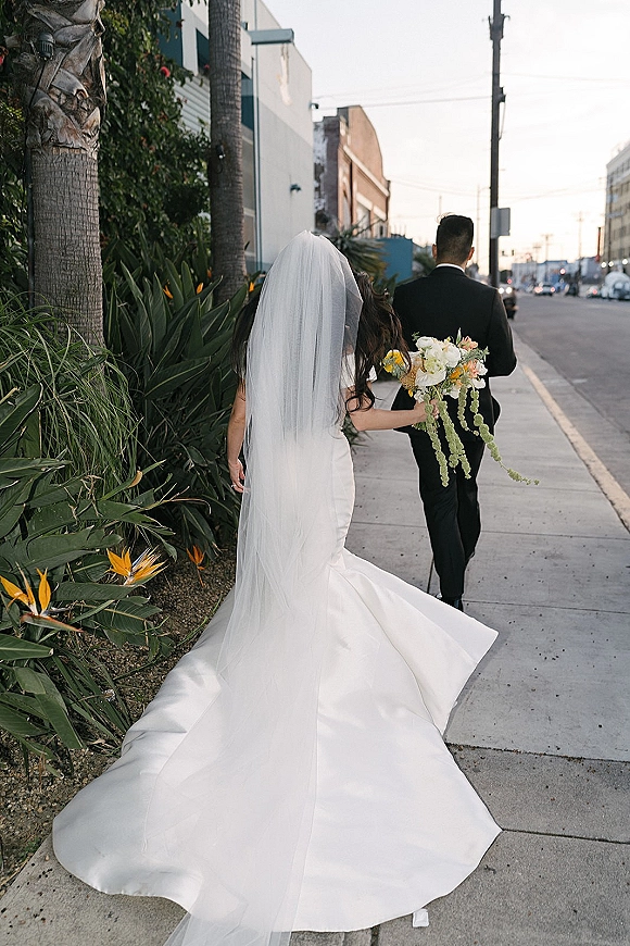 Wedding couple portrait of bride and groom walking away on a city sidewalk, her long veil and dress train flowing, holding a bouquet behind her back