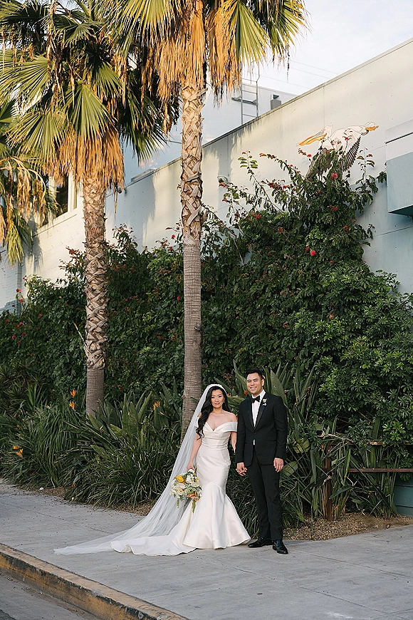 Couple portrait of bride and groom holding hands, bride with bouquet and long veil, groom in black tuxedo by ivy wall and palms