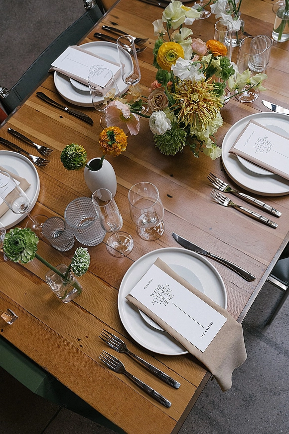 Reception tablescape with wedding table setting on a wood dining table, featuring colorful florals, bud vases, and mixed glassware on concrete floor