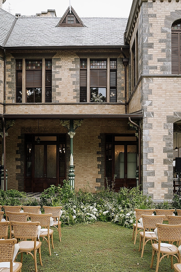 Outdoor ceremony setup with garden wedding ceremony seating, woven chairs flanking a white and greenery floral aisle on a stone manor lawn