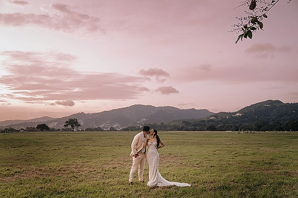 Wedding couple portrait of bride and groom kissing at sunset, her long-train dress and his boutonniere framed by mountains and clouds