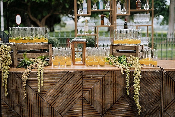 Wedding bar setup with orange cocktails in lined glasses on a rustic wooden bar, liquor shelves, lantern and greenery on a lawn by trees