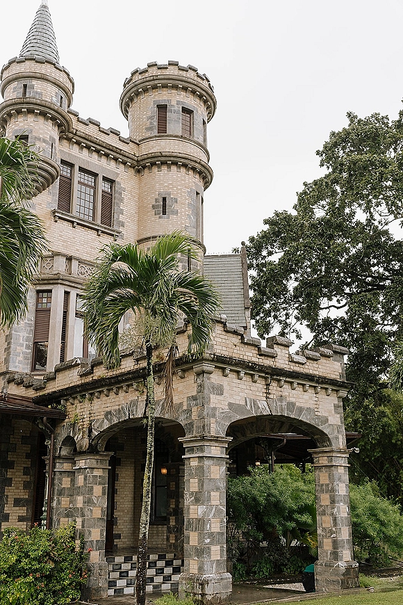 Castle wedding venue exterior with stone arches and turrets, checkered tile steps leading to an arched entryway with palm greenery under overcast sky