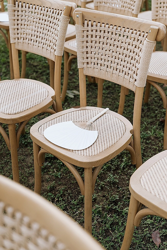Ceremony seating with outdoor ceremony chairs in natural wood, woven cane backs and a hand fan resting on one chair on grass