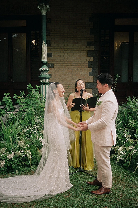 Wedding vows as bride in lace dress and veil holds hands with groom in suit beside mic stand, greenery and brick porch behind