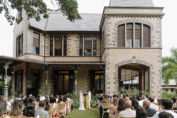 Wedding ceremony with bride and groom at a floral arch, guests in wooden chairs, set before a stone mansion with arched doorway