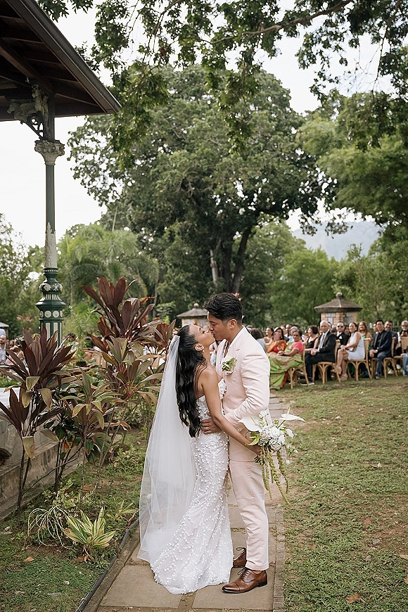 Wedding kiss at an outdoor ceremony as bride in long veil and beaded dress embraces groom in light pink suit on a stone walkway aisle