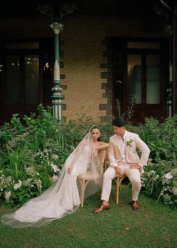 Couple portrait of bride and groom seated on chairs, her long veil draping over a lace wedding dress, in a garden by brick porch columns