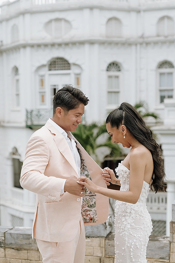 First look moment as bride in a strapless lace wedding dress adjusts groom’s light pink suit by a white facade with palm fronds