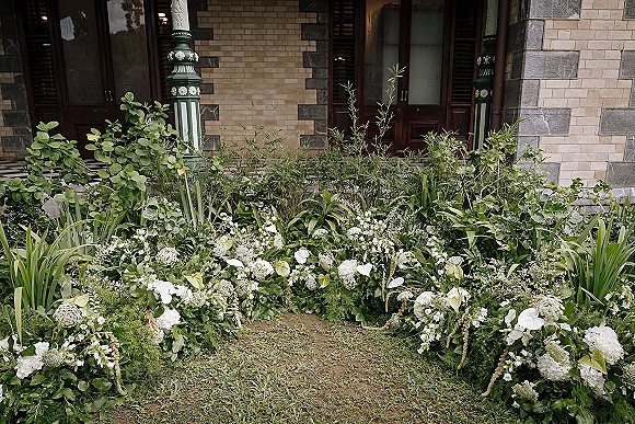 Wedding floral installation with a ground floral arch of white flowers and lush greenery on a lawn before brick doors and columns