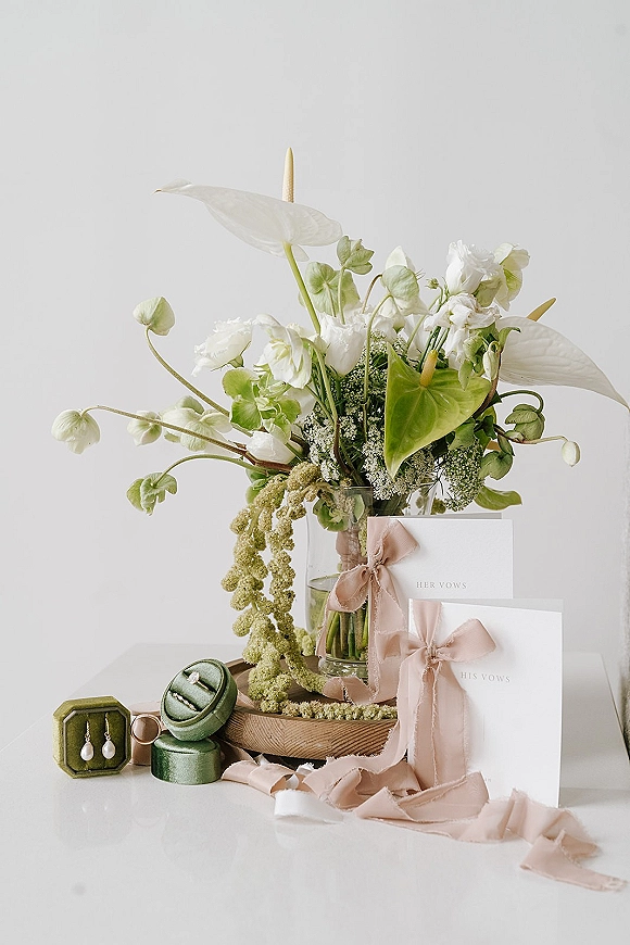 Wedding detail flatlay with a wedding invitation flatlay, vow booklets, rings, pearl drop earrings, silk ribbon, and modern white flowers on a white tabletop