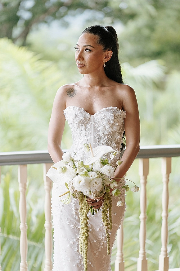 Bridal portrait of a bride in a strapless lace wedding dress holding an all-white anthurium and rose bouquet on a balcony with palms