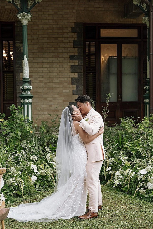 Wedding kiss portrait of bride and groom kissing, her veil flowing over a lace dress train on a garden lawn by a brick porch door