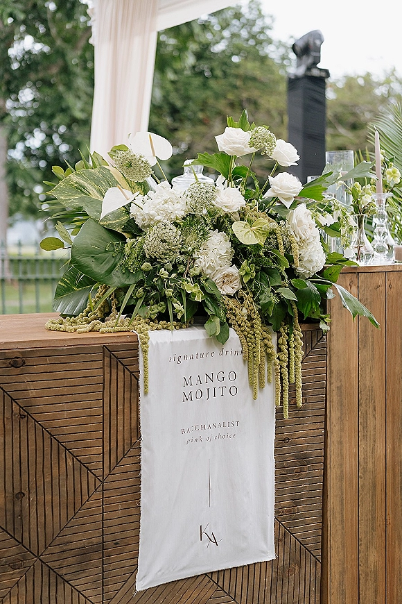 Wedding bar decor with wedding bar signage on a wooden bar, white florals and tropical greenery, glassware, and taper candles outdoors under drapery