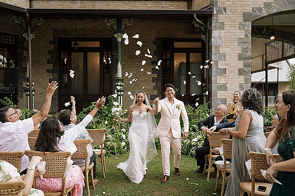 Recessional moment as newlyweds walk down the aisle hand in hand while guests toss petals outside a brick building facade