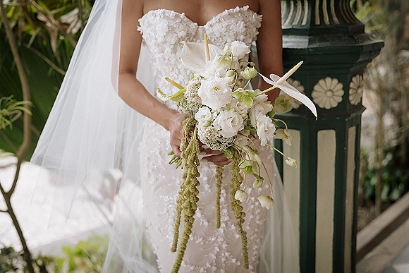 Bridal bouquet with white orchid blooms and anthurium, ribbon and hanging amaranthus, held against a strapless lace dress in garden greenery