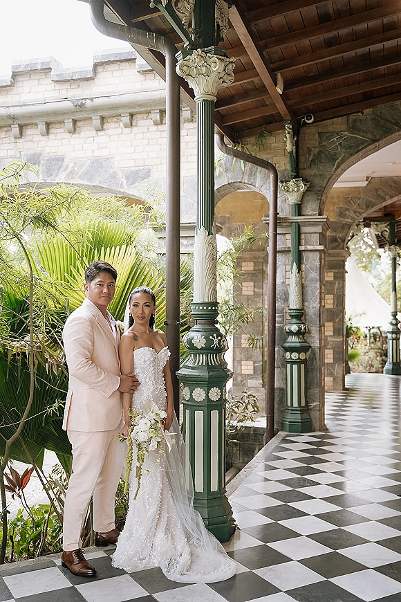 Couple portrait of bride and groom posing on a covered veranda, her lace dress and veil with bouquet, him in beige suit by stone arches