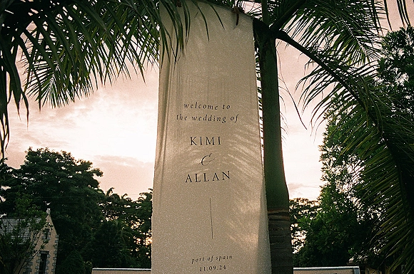 Wedding welcome sign on a hanging fabric banner with printed lettering, framed by palm trees and a sunset sky outside a building