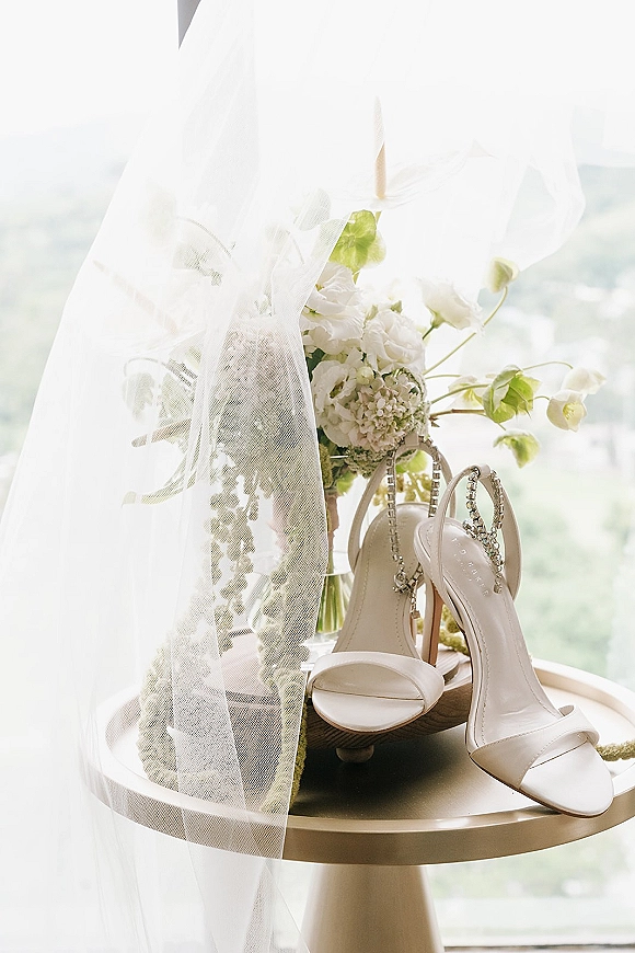 Wedding shoes with bridal heels, white strappy heels and rhinestone ankle straps beside veil and bouquet on a pedestal by window light