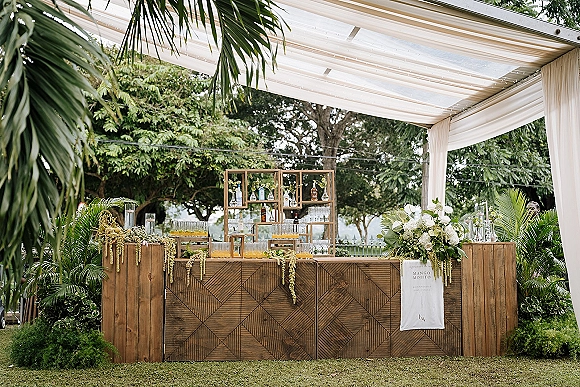 Wedding bar setup with rustic wood facade, glassware, liquor bottles, and cocktail signage under white draping in a garden pergola setting