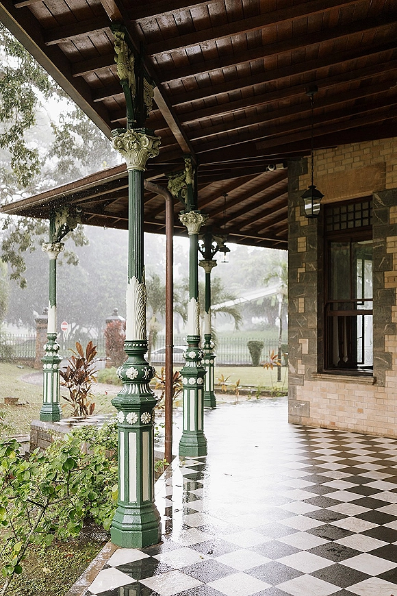 Wedding venue exterior with ornate green columns and a covered porch, black-and-white checkered tile reflecting rain beside garden plants and lawn