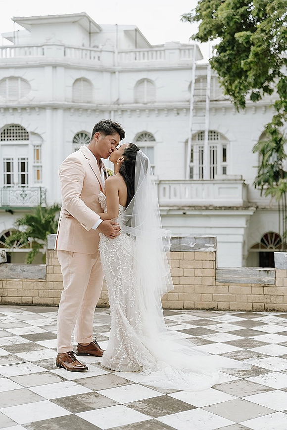 Wedding kiss portrait of bride and groom kissing full-length on a checkered terrace, long veil trailing by a white historic building facade