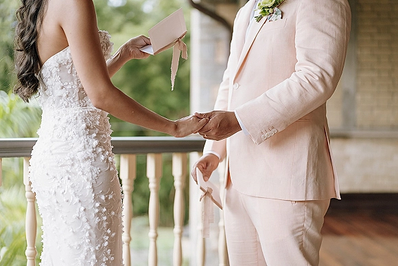 Wedding vows as couple exchange vows holding ribbon-tied vow books, hands together beside lace dress and blush suit on porch railing