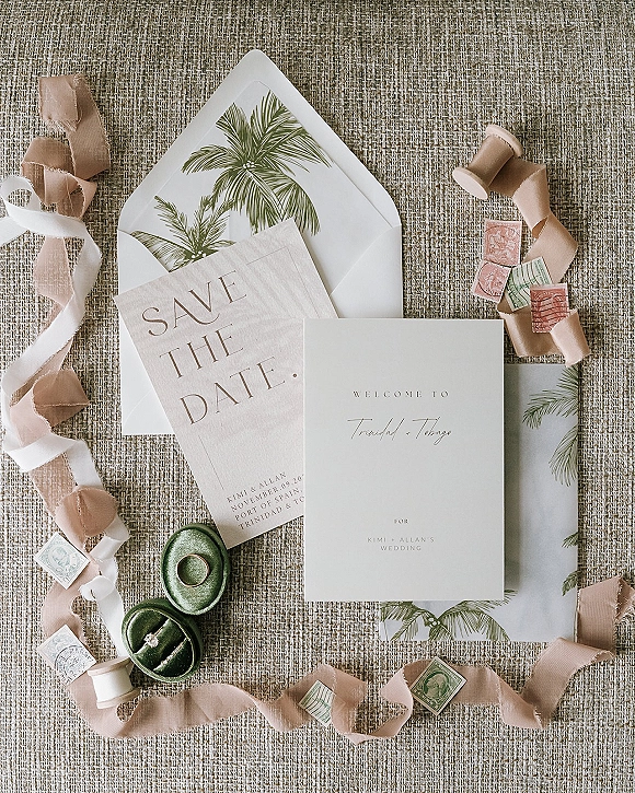 Wedding invitation suite flatlay with save the date card, silk ribbon, vintage postage, and rings on a woven linen backdrop
