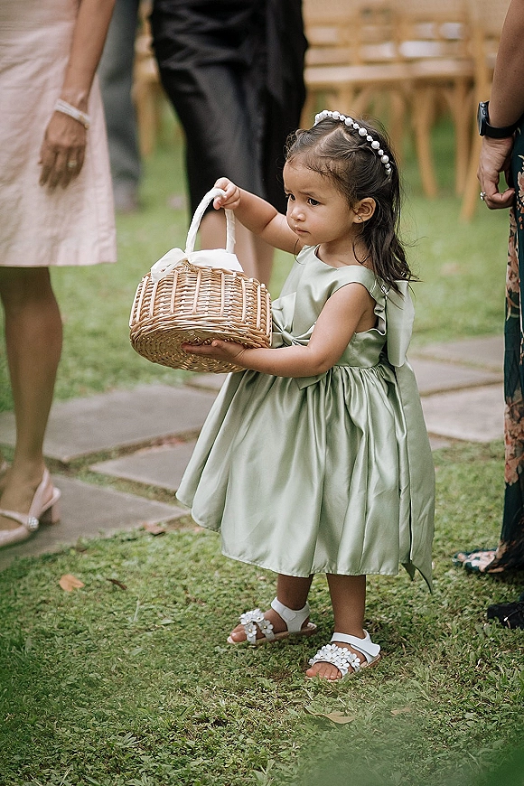 Flower girl holding a flower girl basket in a sage green satin dress with pearl headband, walking on grass by guests and chairs