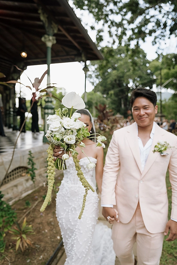 Couple portrait of bride and groom holding hands, bride hiding behind a calla lily bouquet with trailing amaranthus in a garden pavilion with string lights