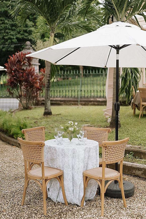 Outdoor reception table with white linen, small floral centerpiece and wine glasses, rattan chairs under a patio umbrella on a gravel patio with palms
