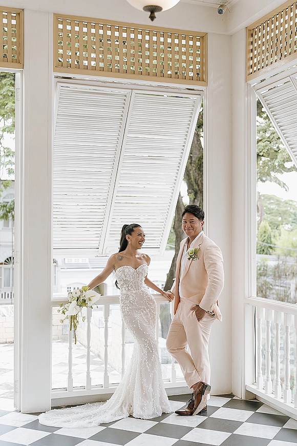 Couple portrait of bride and groom holding hands, laughing on a bright indoor balcony with white shutters and checkered tile floor