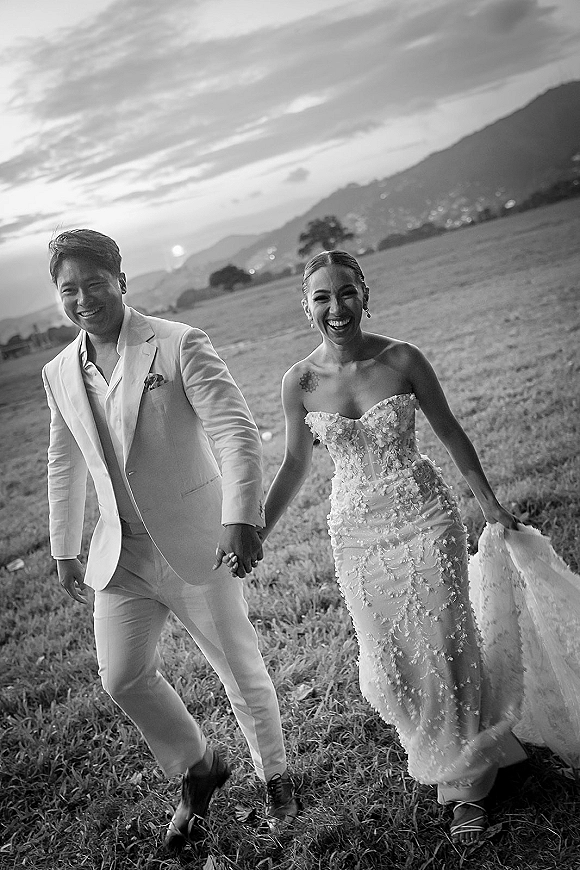 Couple portrait of bride and groom holding hands, walking and laughing in a grassy field with mountains at sunset, her earrings catching light