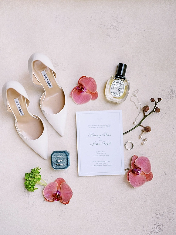 Wedding flat lay of invitation suite with white pointed-toe heels, rings in a box, pearl jewelry, perfume, and orchid stem on stone surface