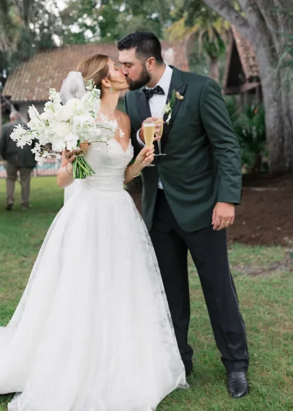 Wedding kiss portrait of bride and groom kissing, holding champagne flutes beside a white bouquet on a garden lawn with trees and tiled-roof building