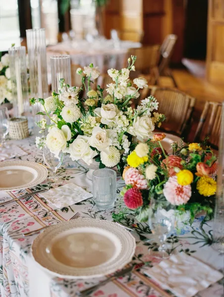 Reception tablescape with wedding table centerpiece, patterned tablecloth, place cards, and ribbed glass votives in an indoor room by windows