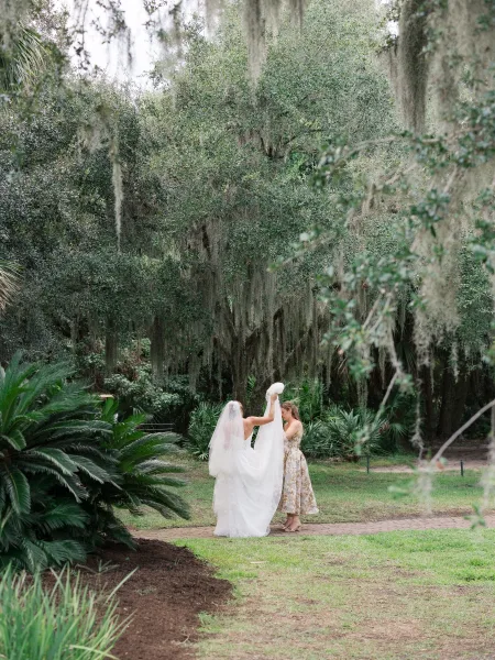 Bridal veil moment as a bride adjusts a long sheer veil over her strapless wedding dress on a brick garden path under trees with spanish moss