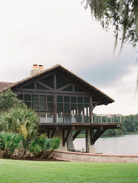Wedding venue exterior with a wraparound deck and lantern lights, stone columns and large windows overlooking a lake with trees and moss under cloudy sky