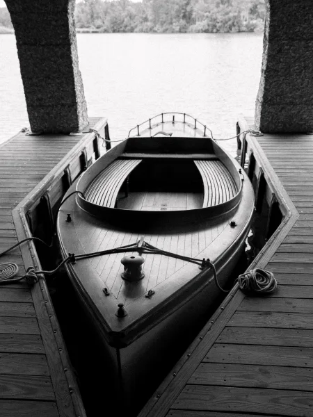 Wooden boat rowboat at dock tied with mooring ropes beside a wooden dock, benches and cleats visible on calm lake near trees