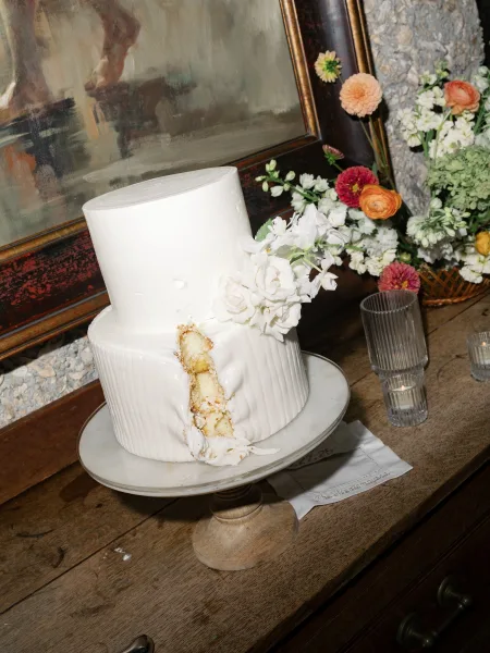 Wedding cake with a two tier wedding cake design, white frosting and floral topper on a stand, styled with ribbed glass votives on a wood table by a stone wall