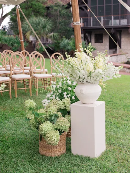 Ceremony florals with an aisle floral arrangement of white blooms and green hydrangeas in baskets, urns, and pedestal plinths on a grassy lawn near wooden chairs