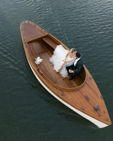 Wedding kiss portrait of bride and groom on boat, her veil and white bouquet beside a wooden boat, floating on calm water