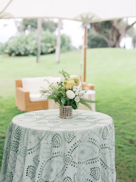 Wedding cocktail table with a floral centerpiece in a woven basket vase on a patterned cloth, set on a lawn under a white tent canopy